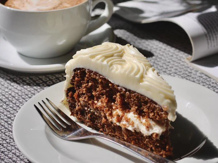 Et stykke gulerodskage med flødeostglasur på en hvid tallerken med en gaffel ved siden af en kop kaffe og en åben bog.