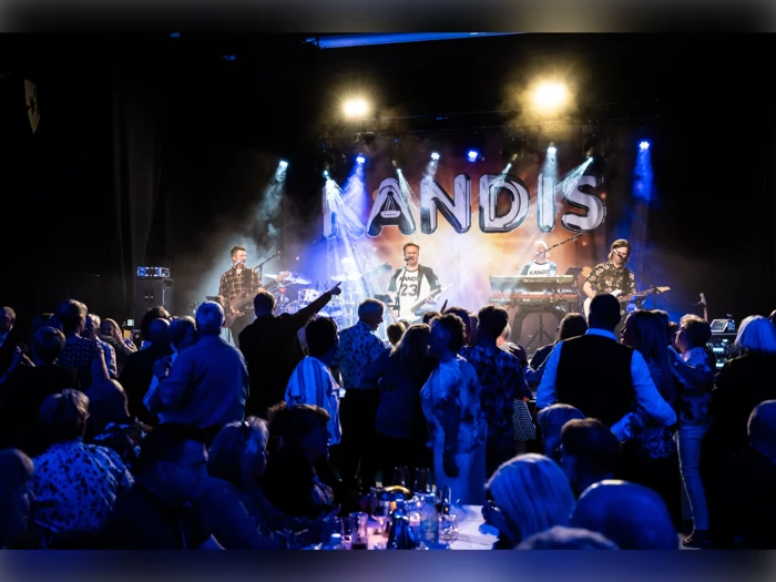 Et liveband optræder på scenen i lyset fra KANDIS, mens publikum ser på, og nogle danser foran scenen.