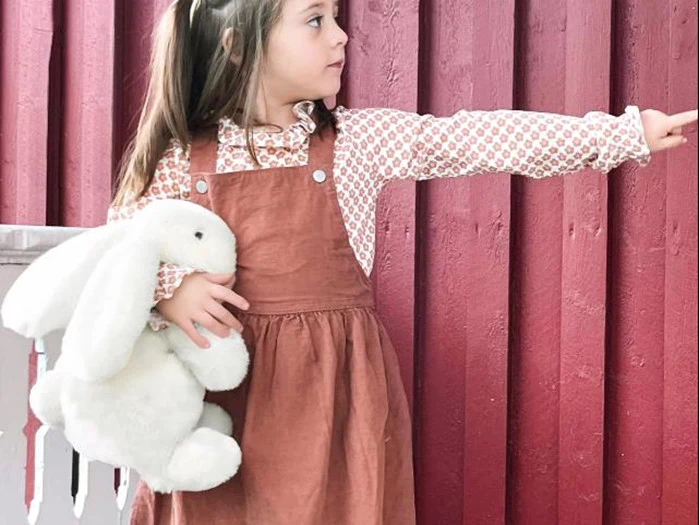 Ung pige i brun kjortel med en hvid kanin i hånden står ved en rød trævæg og peger mod højre.