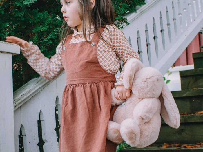 En ung pige i brun kjole holder en stor kanin i plys og står ved en udendørs trætrappe med grønne planter i baggrunden.