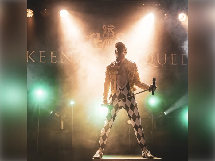 En performer i gul jakke og harlekinbukser står på scenen og holder et mikrofonstativ med dramatisk belysning og røg bag sig.