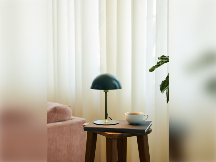 Et lille bord med en sort lampe, en kop kaffe og en bog står ved siden af en lyserød sofa foran hvide gardiner. En grøn plante er delvist synlig til højre.