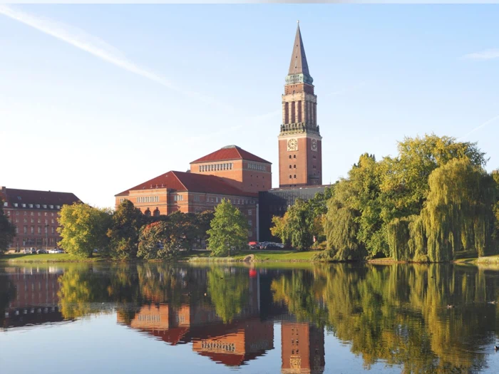 En stor murstensbygning med et højt klokketårn spejler sig i en rolig sø, omgivet af træer under en klar himmel.