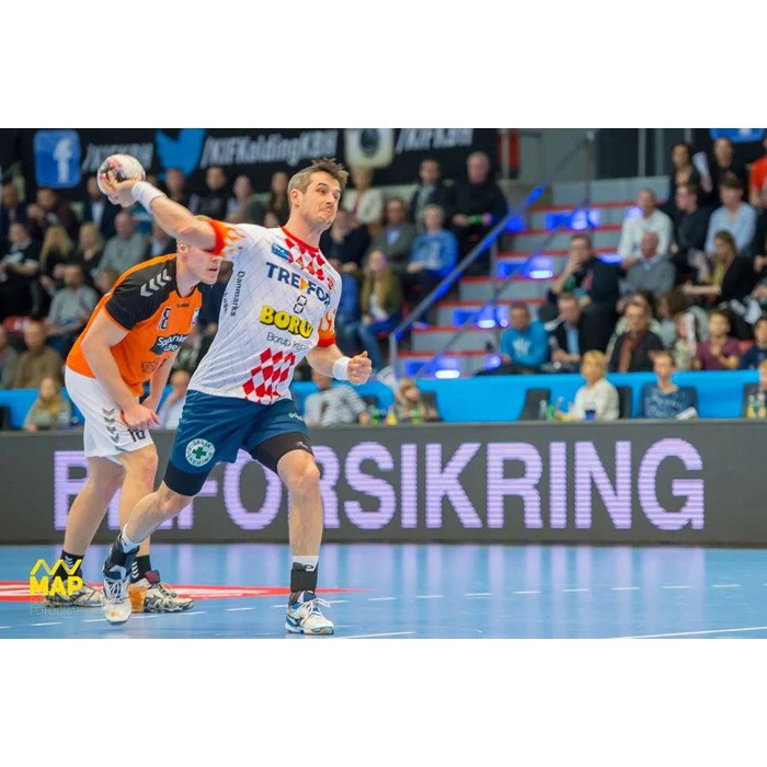 En mandlig håndboldspiller i hvid og rød uniform er ved at kaste bolden, mens en anden spiller i orange uniform står bag ham på en indendørs bane. Der er tilskuere i baggrunden.