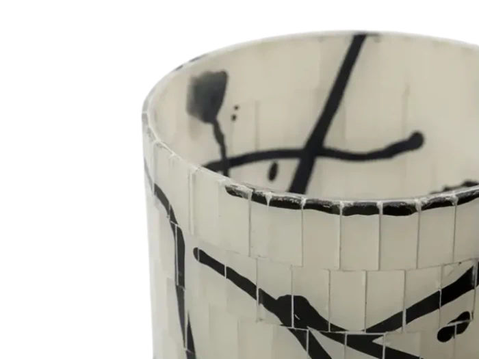 Nærbillede af en cylindrisk vase med hvid mosaikoverflade og abstrakte sorte streger og stænk som dekoration.