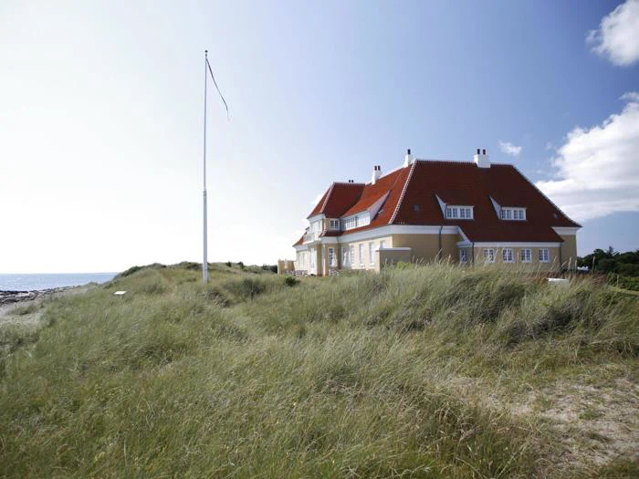 Et stort hus med rødt tag ligger bag højt græs nær en strand under en blå himmel med spredte skyer.