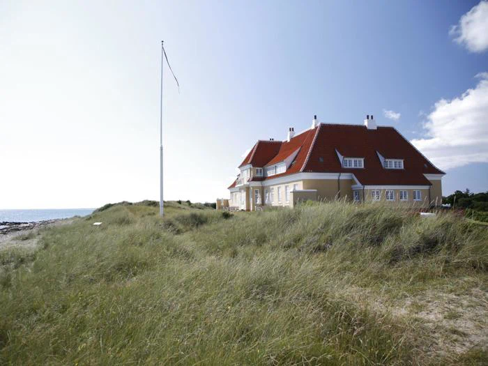 Et stort hus med rødt tag ligger blandt højt græs nær kysten under en klar blå himmel med en tom flagstang foran.