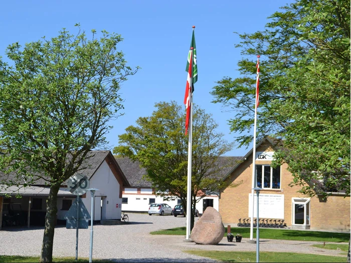 En gårdsplads med bygninger, grønne træer, to flagstænger med flag, en stor sten, et 30 km/t-skilt og flere parkerede biler under en klar blå himmel.
