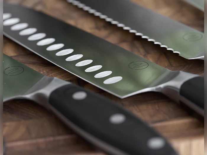 Tre skarpe køkkenknive med sorte håndtag hviler på et skærebræt af træ; den ene kniv har ovale fordybninger på bladet, den anden har en savtakket kant.