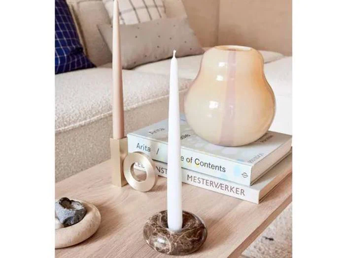 Et moderne sofabord med to bøger, en cremefarvet vase, en rund stenlysestage og to bloklys i forskellige stager. En sofa med puder står i baggrunden.