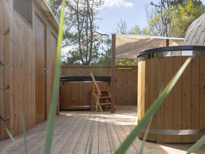 Udendørs træterrasse med et rundt spabad, små trin, en baldakin, et hegn og omgivende træer, set gennem højt græs i forgrunden.