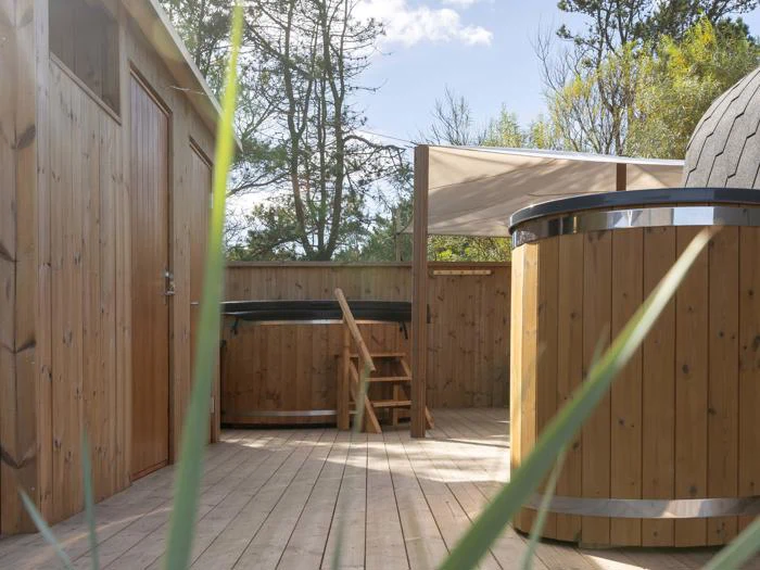 Træterrasse med to runde spabade i træ, en trappe, en træbygning og træer i baggrunden under en delvis overskyet himmel. Græsstrå er synlige i forgrunden.