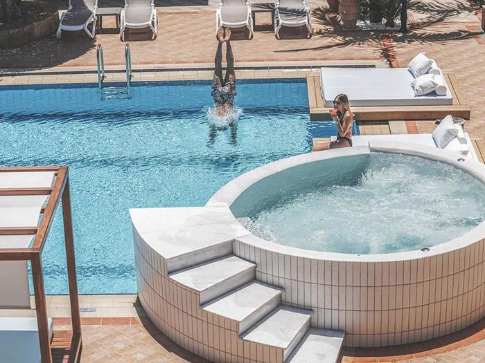 En person dykker ned i en swimmingpool i nærheden af et rundt boblende spabad, mens en anden person sidder i nærheden i en liggestol og bruger en smartphone.