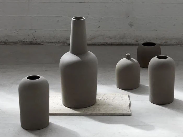 Fem matte, minimalistiske keramikvaser i neutrale toner er arrangeret på et betongulv, og en vase er placeret på en rektangulær stenplade.