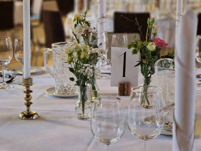 Nærbillede af et elegant dækket bord med glas, små blomsterarrangementer, et hvidt stearinlys og et bordnummer 1-kort på en hvid dug.