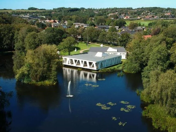 En moderne hvid bygning omgivet af træer ligger ved en dam med et springvand, med en lille by og grønne marker i baggrunden.