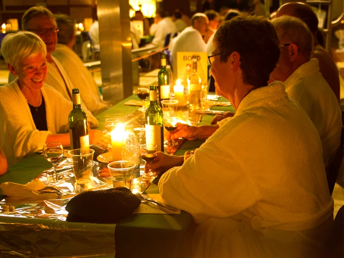 En gruppe ældre i morgenkåbe sidder omkring et bord med stearinlys, vin og mad, smiler og nyder en varm, hyggelig middag sammen.