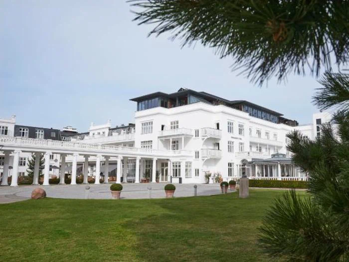 En stor, elegant hvid bygning med balkoner og søjler, omgivet af grønt græs og træer, under en klar himmel.