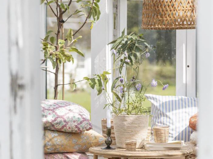 En hyggelig udestue med et lille bord, potteblomster, stablede puder, en kurvelampe og sollys, der strømmer ind gennem store vinduer.