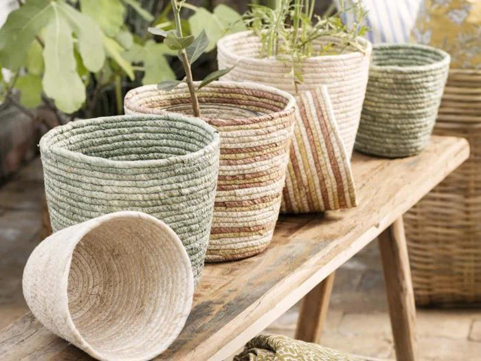Fem flettede kurve i jordfarver, nogle med planter, er arrangeret på en rustik træbænk udendørs. En kurv er væltet om på siden i forgrunden.