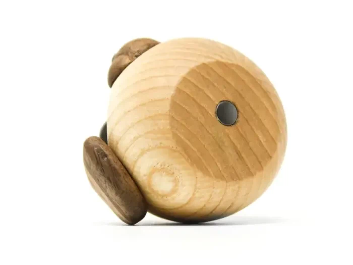En rund træfigur med mørkere træstykker til ører og fødder, der ligner et minimalistisk dyr, på en hvid baggrund.