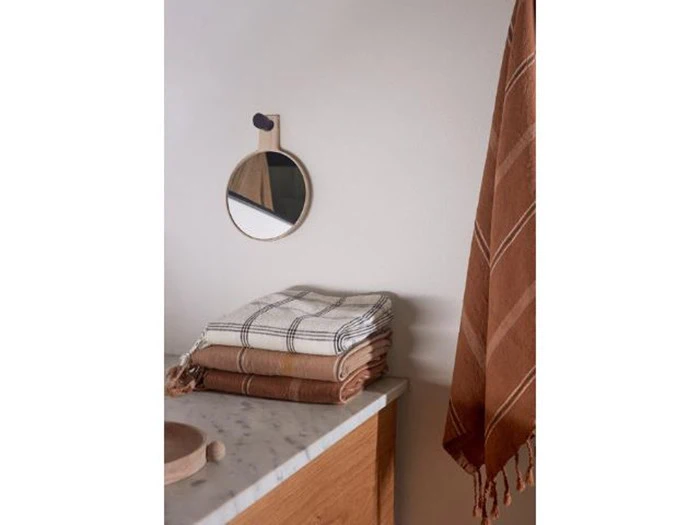 Et lille rundt spejl på en væg over en marmorbordplade med foldede ternede håndklæder og et brunt håndklæde hængende ved siden af.
