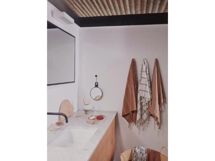 Moderne badeværelse med marmorbordplade, trædetaljer og tre håndklæder hængende på væggen. Et rundt spejl og en kurv er også synlige. Loftet har synlige træbjælker.