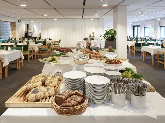 Et buffetbord med tallerkener, bestik, brød, frugt og salater i et rummeligt spiseområde med pænt opstillede borde og stole.