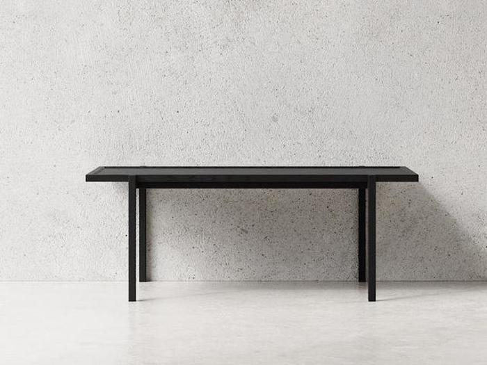 Et minimalistisk sort rektangulært bord med rene linjer står mod en almindelig lysegrå struktureret væg og et glat hvidt gulv.