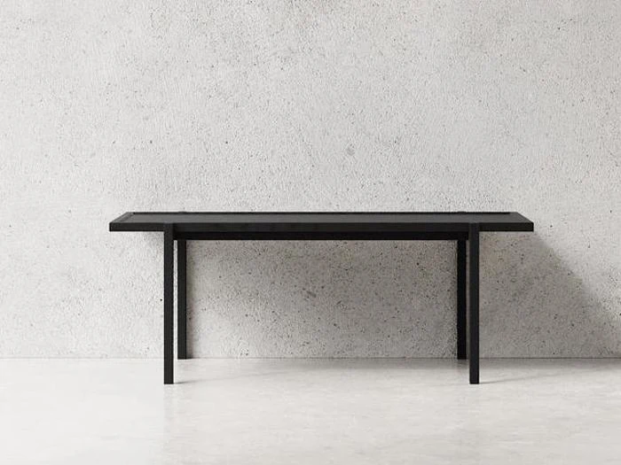 Et minimalistisk sort rektangulært bord med rene linjer står mod en almindelig lysegrå struktureret væg og et glat hvidt gulv.