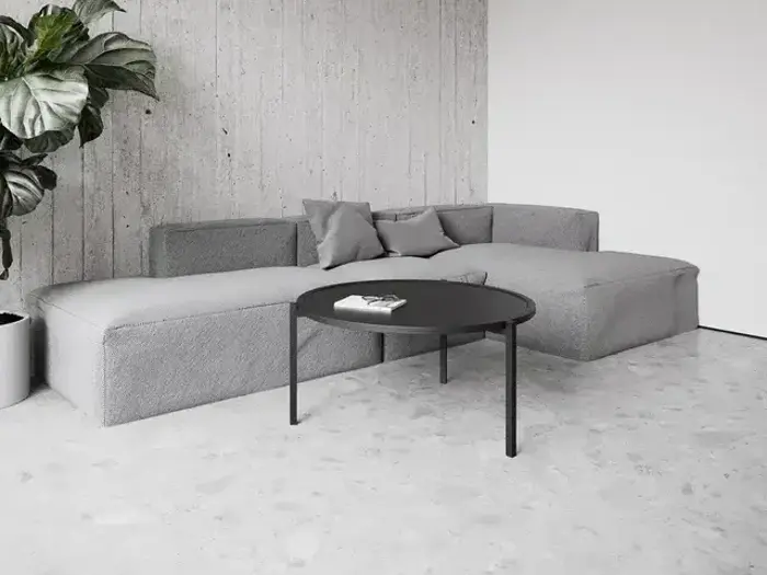 En moderne stue med en grå sektionssofa, et sort rundt sofabord, minimalistisk indretning og en stor potteplante i hjørnet.