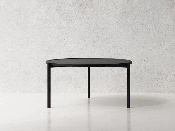 Et rundt, sort bord med fire ben står på et lyst gulv mod en ensfarvet, struktureret grå væg.