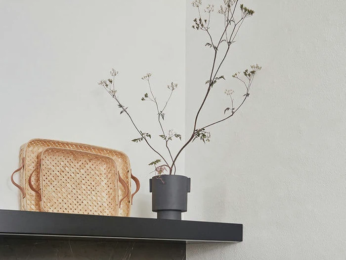 En høj, tynd gren med små hvide blomster i en sort vase står ved siden af to flettede kurve på en sort hylde mod en hvid væg.