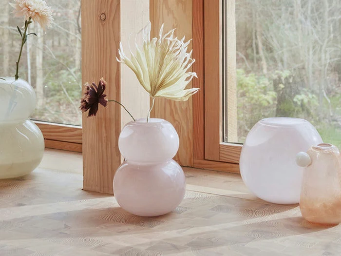 Runde glasvaser i pastelfarver med et par blomster er placeret i en vindueskarm af træ ved siden af et vindue med udsigt til en have.