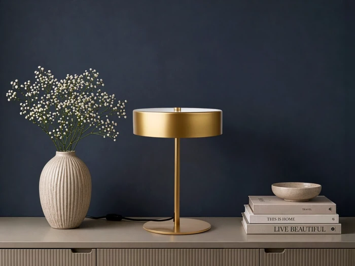 En guldfarvet bordlampe står på et beige skænk ved siden af en vase med hvide blomster, en stak bøger og en lille skål, med en mørkeblå væg i baggrunden.