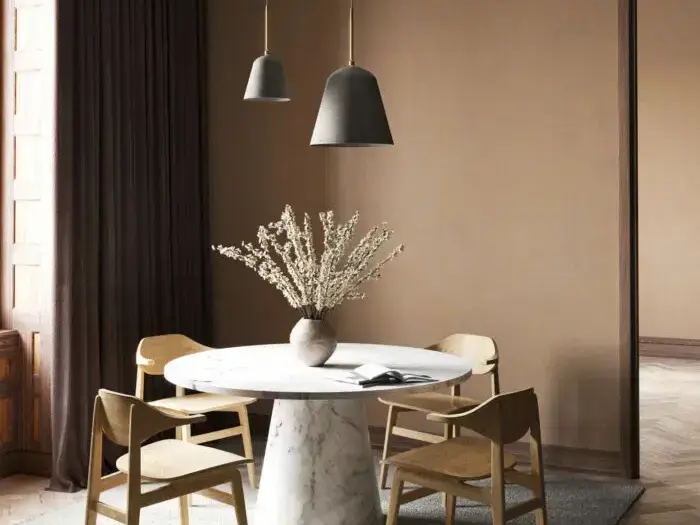 Et rundt hvidt marmorbord med en vase med tørrede blomster er omgivet af fire træstole i en minimalistisk spiseplads med beige vægge og to pendellamper.