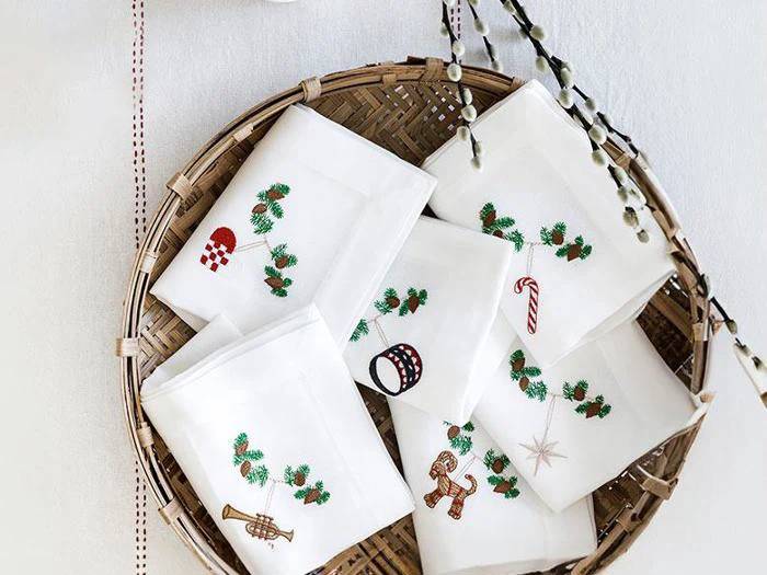 En flettet kurv rummer syv hvide servietter, som hver især er dekoreret med festlige broderede motiver som en sukkerstang, en honningkagemand, en tromme, fyrretræsgrene og julepynt.