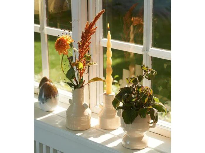 Tre cremefarvede vaser står i en solrig vindueskarm; den ene indeholder blomster, den anden et snoet stearinlys og den tredje en bladplante. En reflekterende æggeformet genstand er også til stede.