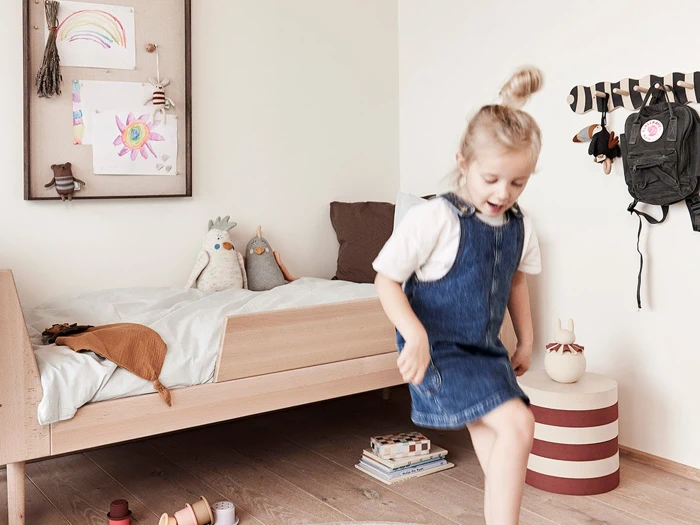 Et lille barn i denimkjole leger i et hyggeligt soveværelse med en træseng, plyslegetøj, vægkunst og rygsække, der hænger på kroge. Legetøj og bøger ligger spredt på trægulvet.