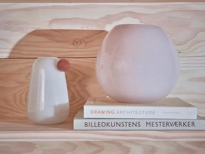 To moderne keramikvaser står på en træhylde ved siden af tre stablede bøger, heriblandt en med titlen "DRAWING ARCHITECTURE". Baggrunden består af lyse træpaneler.