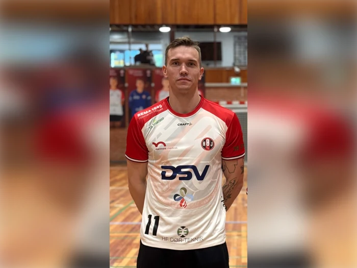 En mandlig håndboldspiller iført en hvid og rød trøje med nummer 11 og forskellige sponsorlogoer står i en idrætshal.