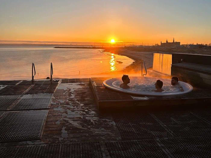 Tre personer slapper af i et spabad med udsigt til en rolig strand ved solnedgang, hvor det orange lys reflekteres i vandet og fjerne bygninger i horisonten.
