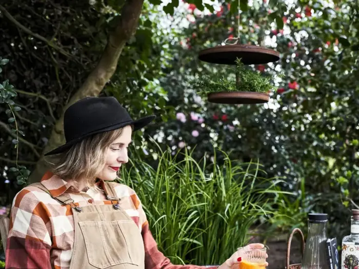 En kvinde i ternet skjorte og solbrændt overall med sort hat holder en drink, mens hun sidder udendørs i nærheden af grønne planter og en hængende krukke.