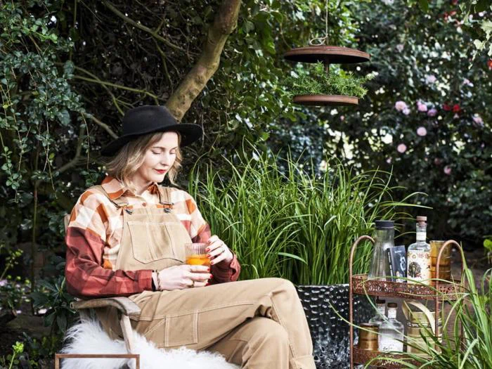 En person i overalls og hat sidder i en have med en drink i hånden. Højt græs, træer og en drinksvogn med flasker og glas er omkring dem og skaber et fredeligt udendørs miljø.