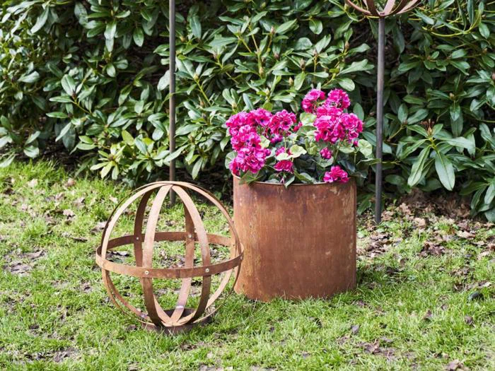 En rusten metalplante med lyserøde blomster står på græs ved siden af en dekorativ havekugle i metal med grønne buske i baggrunden.
