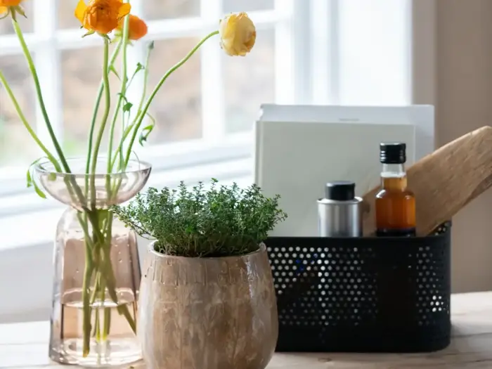 På et træbord står en glasvase med gule blomster, en potteplante og en sort kurv med flasker, papirer og et træredskab ved siden af et lyst vindue.