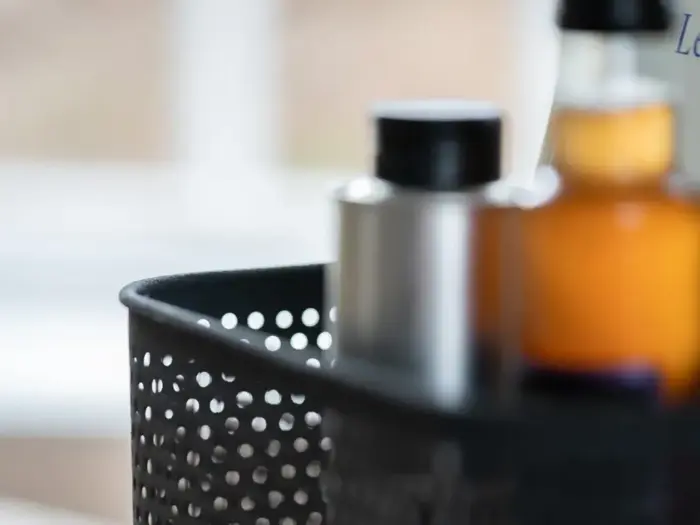 Nærbillede af en sort, perforeret kurv med flasker, bl.a. en sølvflaske og en lille ravfarvet flaske, med sløret baggrund.