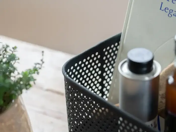 En sort perforeret kurv rummer en metaldåse og en bog; en grøn potteplante står på et træbord i nærheden.