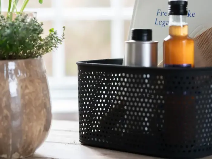 En sort perforeret kurv med flasker står på et bord ved siden af en potteplante, med et vindue og en sløret baggrund bag dem.