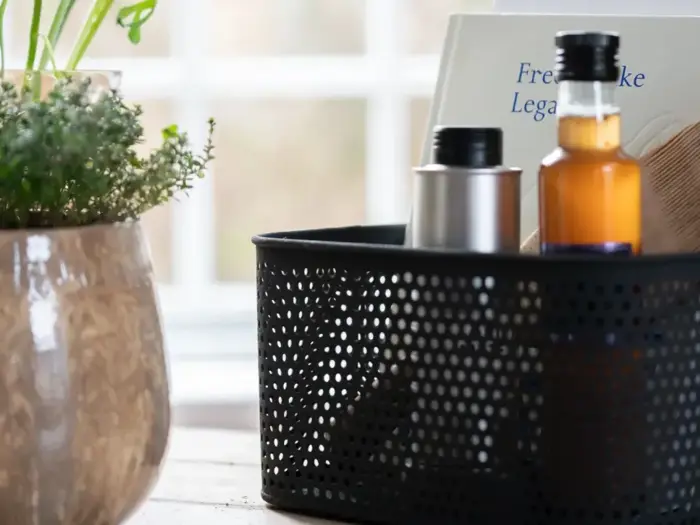 En sort perforeret kurv rummer flasker og en bog ved siden af en potteplante på et lyst bord nær et vindue.